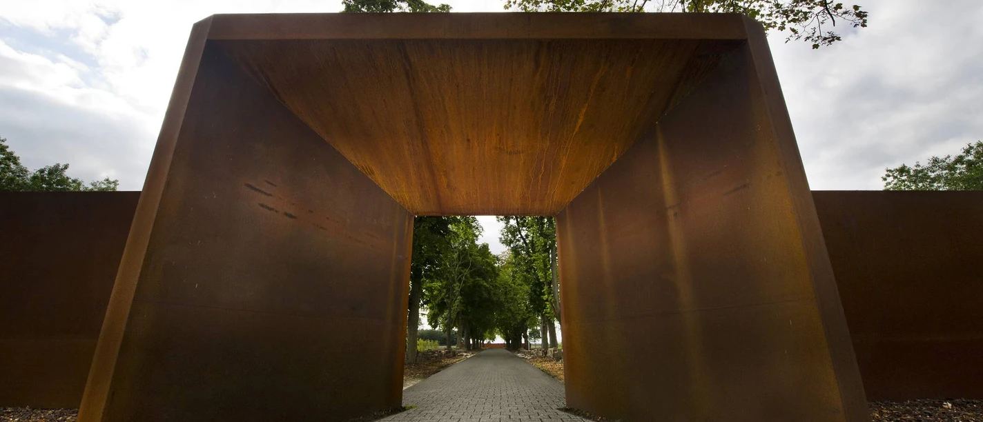 Gedenkstätte Esterwegen Rostfarbener Stahlbogen mit gepflastertem Weg, der in eine von Bäumen gesäumte Allee führt.