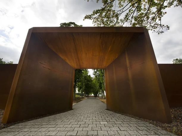 Gedenkstätte Esterwegen Rostfarbener Stahlbogen mit gepflastertem Weg, der in eine von Bäumen gesäumte Allee führt.