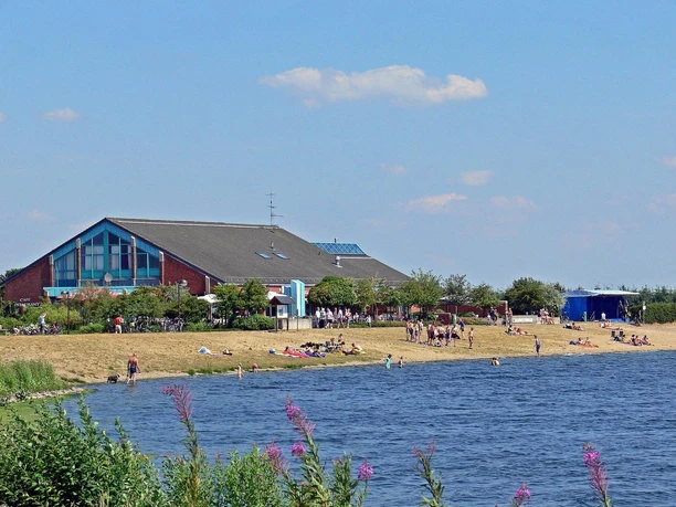 Touristikbüro der Gemeinde Geeste Menschen entspannen am Seeufer vor einem großen roten Gebäude mit Café und blühenden Pflanzen.