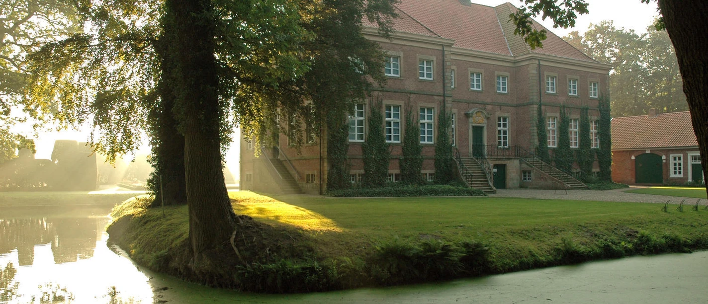 Gut Altenkamp - Überregionales Ausstellungszentrum Historisches Herrenhaus aus rotem Backstein mit Wassergraben, Bäumen und Sonnenlicht im Park.