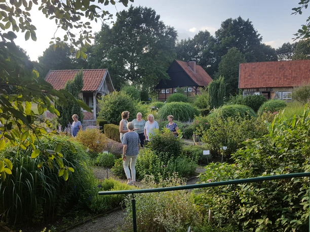 Heilkräutergarten Emsbüren Menschen spazieren durch einen blühenden Kräutergarten mit alten Fachwerkhäusern im Hintergrund.