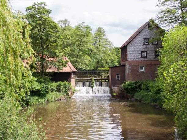 Herrenmühle Meppen Historische Wassermühle aus Backstein mit Holzschindeln an einem kleinen Wasserlauf im Grünen.