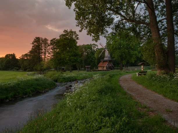 Hüvener Mühle Historische Hüvener Mühle mit Wasserrad und Windflügeln an einem Bach bei Sonnenuntergang im Grünen