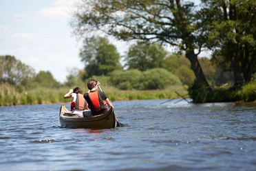 Kanu-Verleih Gasthaus Dröge-Polle Zwei Personen paddeln in einem Kanu auf einem ruhigen Fluss, umgeben von grüner Uferlandschaft.