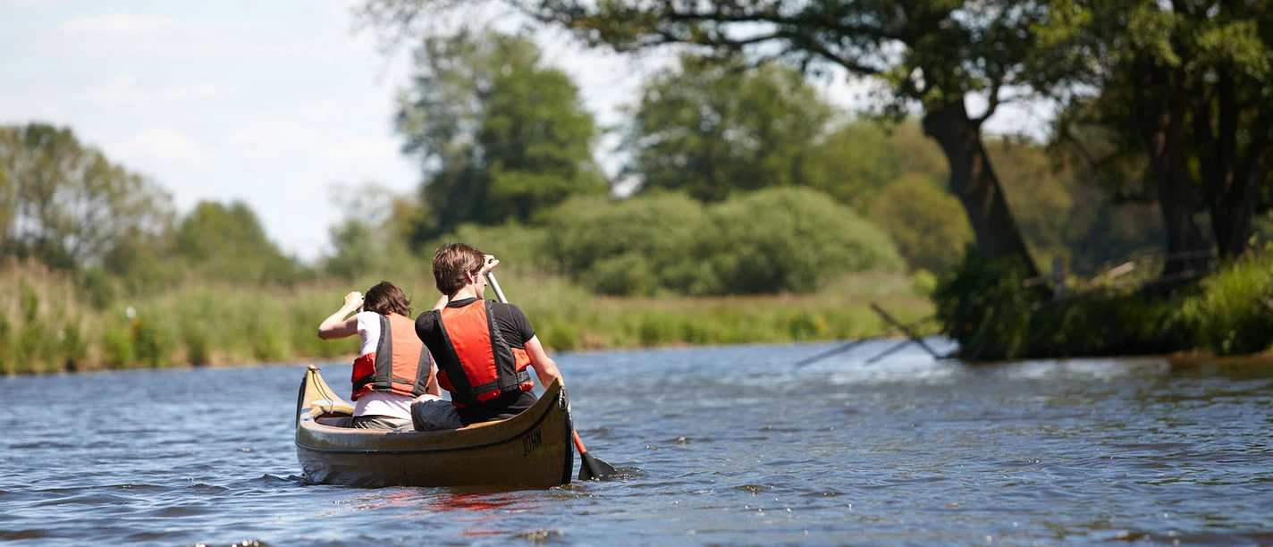 Kanu-Verleih Gasthaus Dröge-Polle Zwei Personen paddeln in einem Kanu auf einem ruhigen Fluss, umgeben von grüner Uferlandschaft.