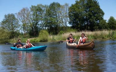 Kanuverleih Hasetal Vier Personen paddeln in zwei Kanus über einen ruhigen Fluss, umgeben von grünem Ufer und Bäumen.