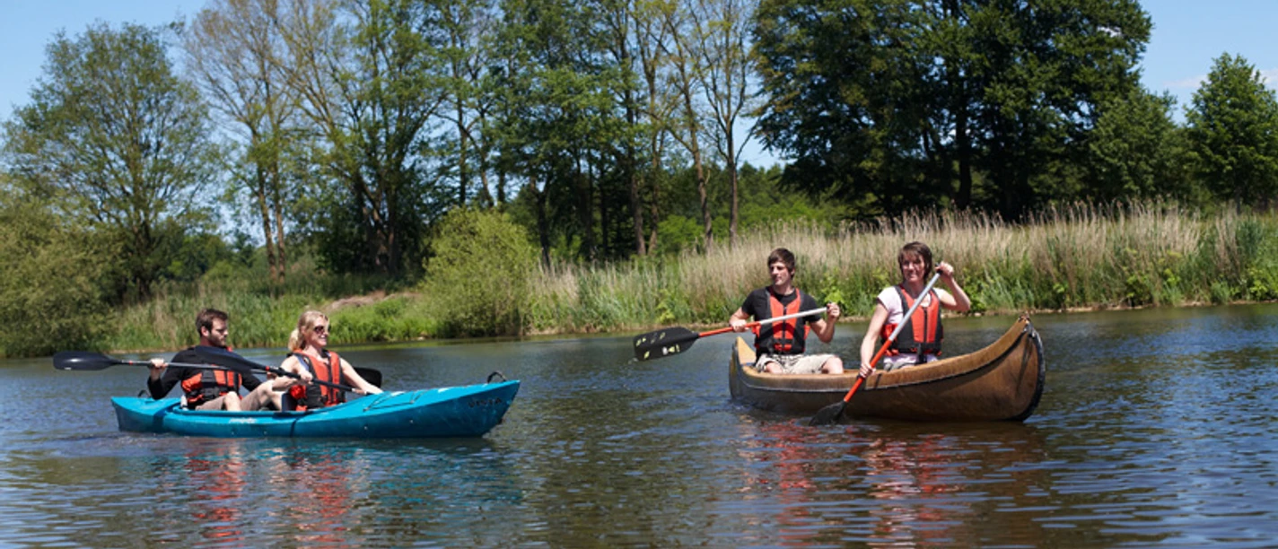 Kanuverleih Hasetal Vier Personen paddeln in zwei Kanus über einen ruhigen Fluss, umgeben von grünem Ufer und Bäumen.