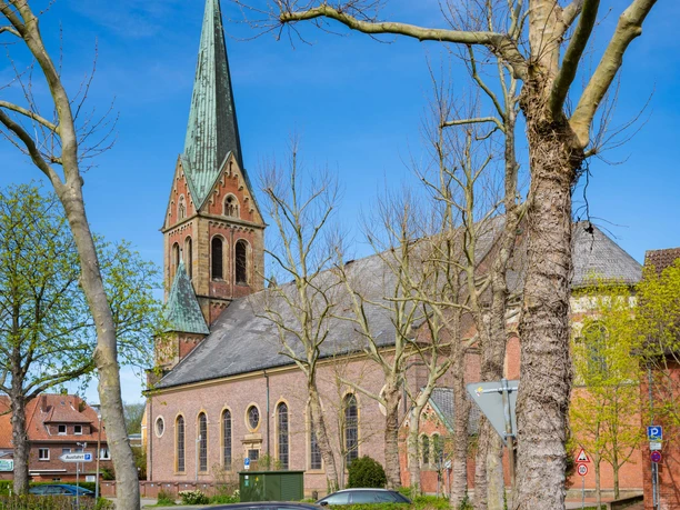 St.-Bonifatius-Kirche in Lingen Backsteinkirche mit hohem Turm und grünem Spitzdach, umgeben von Bäumen und parkenden Autos.