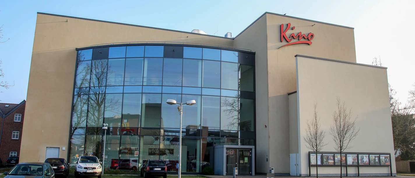 Modernes Kinohaus mit großer Glasfassade, rotem Schriftzug und kleinem Parkplatz im Vordergrund.
