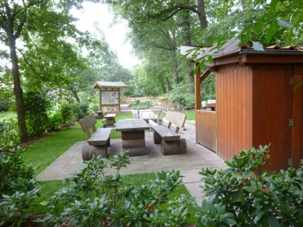 Kneipp-Erlebnisse an der Koppelschleuse Holzhaus mit Sitzgruppe im Grünen neben einer Wassertretanlage unter schattigen Bäumen.