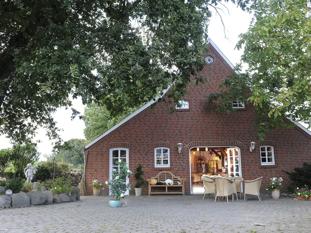 Lotos Salzgrotte Backsteingebäude mit blühenden Pflanzen, Sitzbereich und großen schattenspendenden Bäumen.