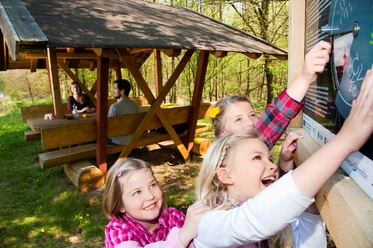 Löwenzahnentdeckertouren Fullener Wald Kinder entdecken fröhlich eine Mitmachtafel im Wald, während Erwachsene an einem Picknickplatz sitzen.