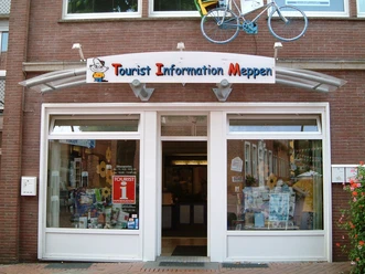 De entree van het VVV-kantoor in Meppen met een glazen pui, informatiemateriaal en een gele fiets boven het bord.