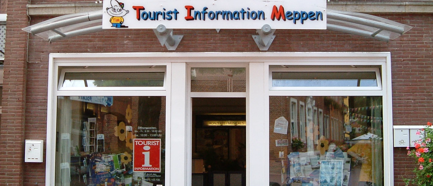De entree van het VVV-kantoor in Meppen met een glazen pui, informatiemateriaal en een gele fiets boven het bord.