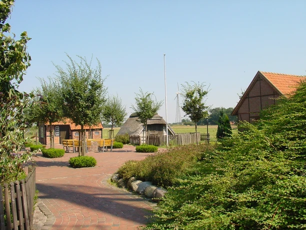 Mühlenhof und Pfarrgarten Wippingen Backsteinhäuser mit roten Dächern, gepflegter Gartenweg, grüne Büsche und Bäume im Sonnenschein.