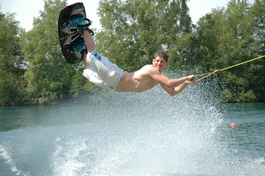 Naherholungsgebiet Heeder See Mann beim Wakeboarden über dem Wasser, spritzendes Wasser, sonniger Tag, See mit grünen Bäumen.