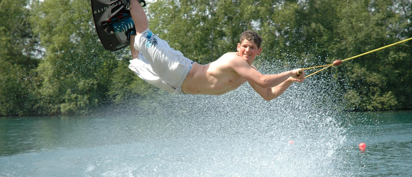 Naherholungsgebiet Heeder See Mann beim Wakeboarden über dem Wasser, spritzendes Wasser, sonniger Tag, See mit grünen Bäumen.