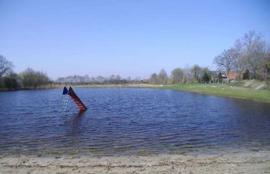 Naturbad Neurhede Kleiner Badesee mit Sandstrand und Wasserrutsche, umgeben von Wiese und Bäumen unter blauem Himmel.