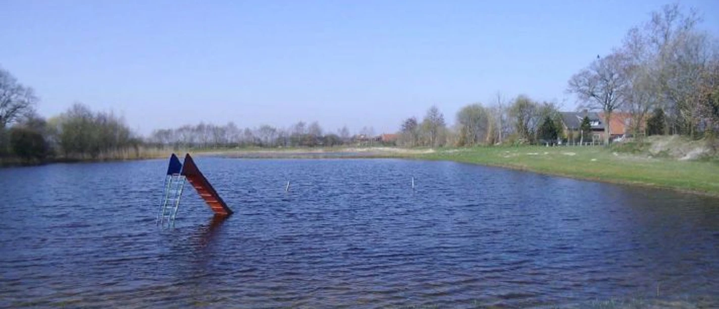 Naturbad Neurhede Kleiner Badesee mit Sandstrand und Wasserrutsche, umgeben von Wiese und Bäumen unter blauem Himmel.