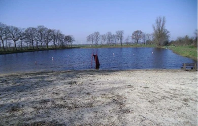 Natursee Brual Kleiner Badesee mit sandigem Ufer, roter Rutsche im Wasser und Bäumen im Hintergrund.