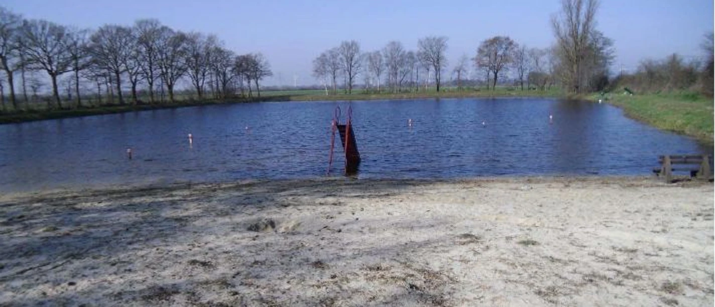 Natursee Brual Kleiner Badesee mit sandigem Ufer, roter Rutsche im Wasser und Bäumen im Hintergrund.