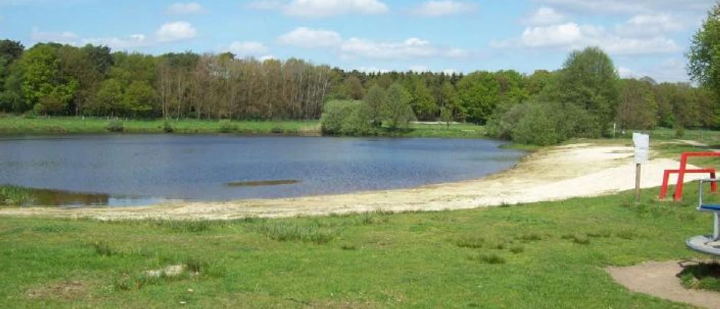 Natursee Campingplatz Grüne Wiese mit Blick auf einen kleinen See, hellem Sandstrand und umgebendem Waldgebiet.