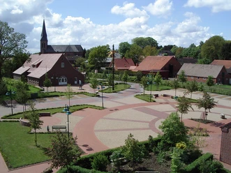 Infozentrum Esterwegen Großzügiger Dorfplatz in Esterwegen mit rotem Klinkerbau, Kirche im Hintergrund und grünen Anlagen.