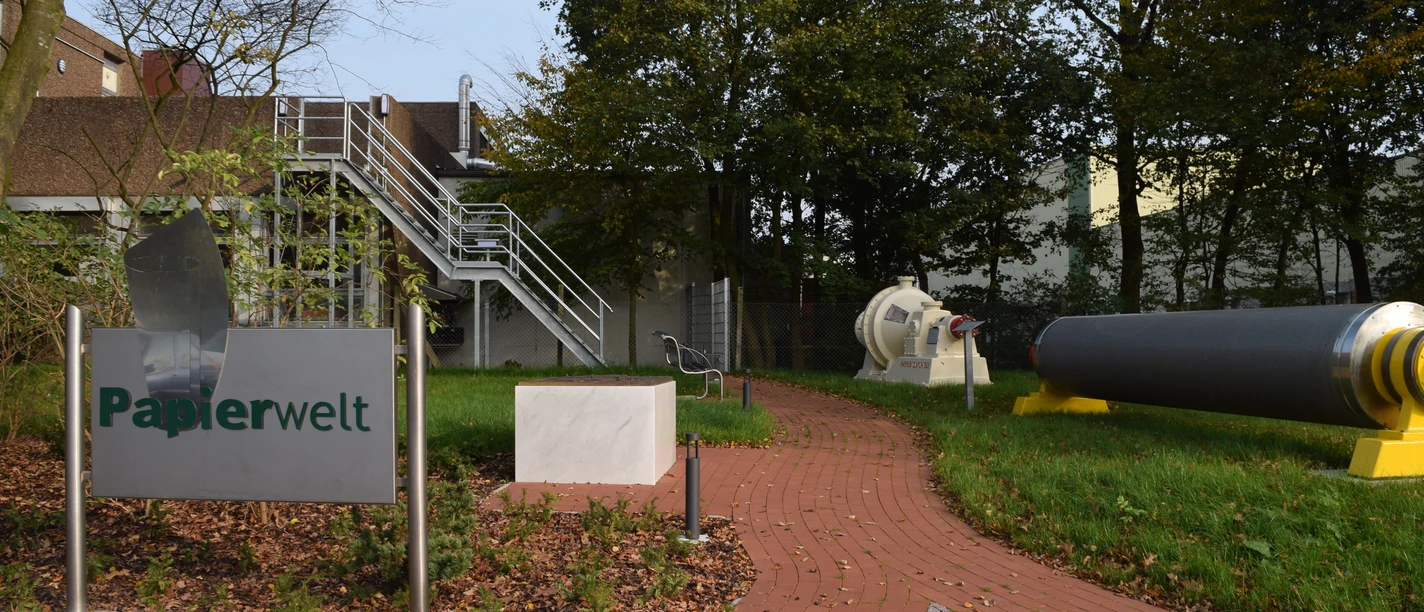 Papierwelt Dörpen Eingangsbereich der Papierwelt Dörpen mit Infotafel, rotem Weg und großen Papiermaschinen-Teilen.