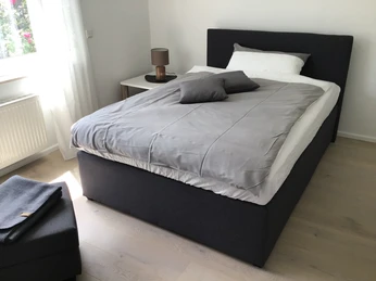 Tannenweg_Schlafen1.1_SHS.JPG Helles Schlafzimmer mit einem Einzelbett, grauer Bettwäsche, Lampe auf dem Nachttisch und großem Fenster.