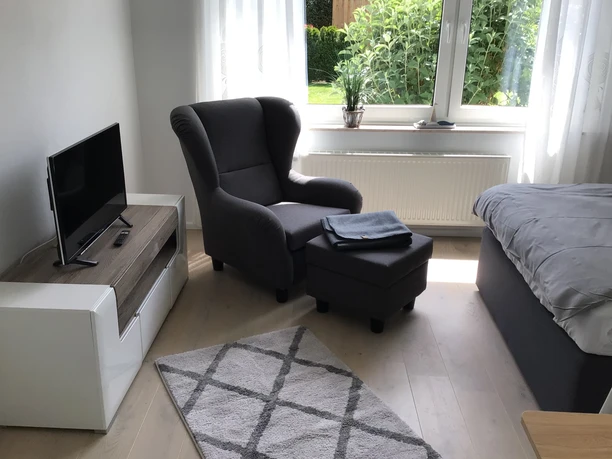 Helles Schlafzimmer mit grauem Sessel und Ottomane, Bett und TV, daneben großes Fenster mit Gartenblick.