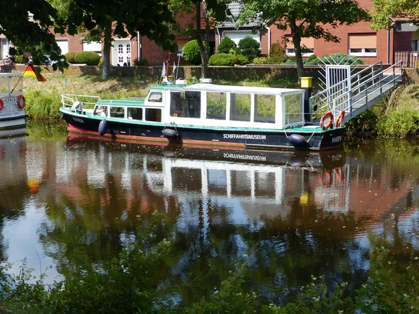 Schifffahrtsmuseum Haren (Ems) Ausflugsboot des Schifffahrtsmuseums Haren liegt am Kanal, umgeben von Bäumen und Backsteinhäusern.