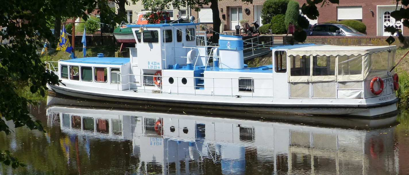 Weißes Museumsschiff mit blauen Akzenten liegt ruhig am Kanal, umgeben von Bäumen und Häusern.