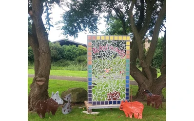 Schöpfungsweg im Burgpark Venhaus Bunte Mosaiktafel zwischen Bäumen mit Tierfiguren aus Metall und Ton auf grünem Rasen.