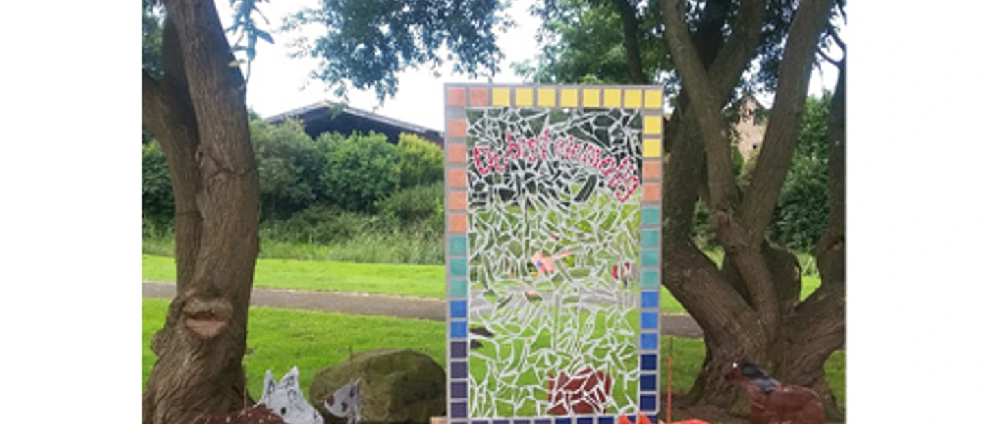 Schöpfungsweg im Burgpark Venhaus Bunte Mosaiktafel zwischen Bäumen mit Tierfiguren aus Metall und Ton auf grünem Rasen.