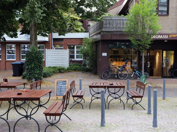 Holzbänke und -tische vor einem Café auf einem gepflasterten Platz mit Bäumen und angrenzendem Gebäude.