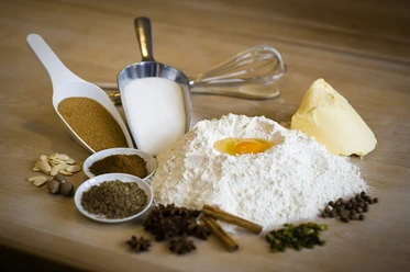 Schulbäckerei Coppenrath Feingebäck GmbH Backzutaten auf Holztisch mit Mehl, Ei, Butter, Zucker, Gewürzen, Nüssen und Backutensilien arrangiert.