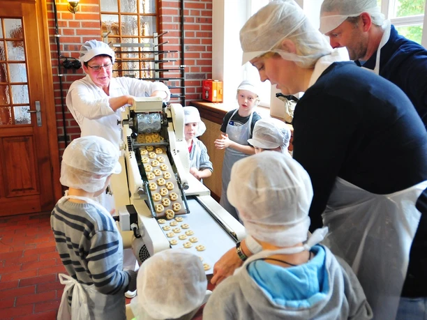 Schulbäckerei Coppenrath Feingebäck GmbH Kinder und Erwachsene formen gemeinsam Gebäck in einer Backstube mit Bäckerin und Maschine.