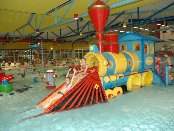 Spaßbad Topas Bunte Spielzug-Lokomotive mit Wasserrutsche in einem belebten Hallenbad voller planschender Kinder.