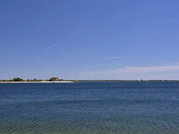 Speichersee Geeste Blauer See mit klarem Wasser unter wolkenlosem Himmel, Ufer mit Strandhaus und Windrädern in der Ferne.