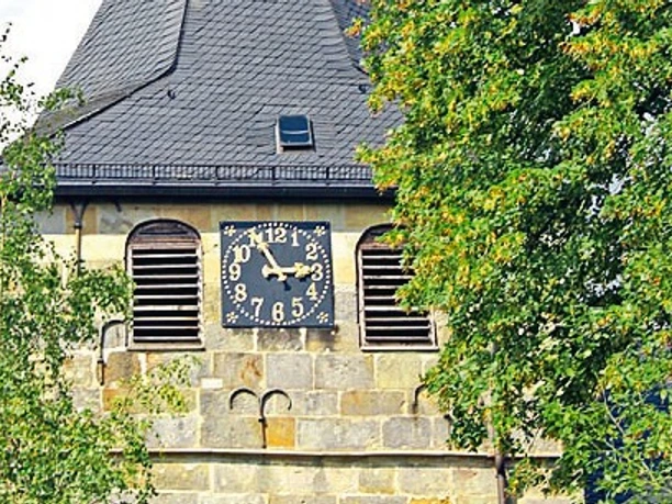 St.-Vitus-Kirche in Bokeloh Turm der St.-Vitus-Kirche in Bokeloh mit schwarzer Turmuhr und Schieferdach hinter grünem Laub.