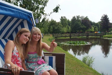 Stadtpark Papenburg Zwei junge Frauen sitzen im Strandkorb am Teich, lächeln und blicken auf eine Brücke im Stadtpark.