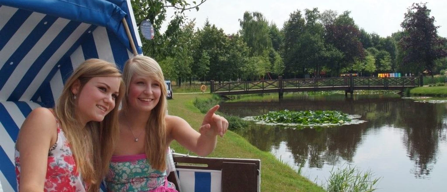 Stadtpark Papenburg Zwei junge Frauen sitzen im Strandkorb am Teich, lächeln und blicken auf eine Brücke im Stadtpark.