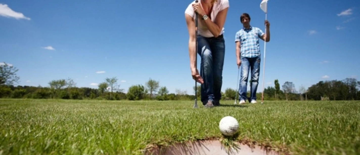 SwinGolf Mehringer Heide Frau beim SwinGolf-Abschlag auf grüner Wiese unter blauem Himmel, Mann mit Flagge im Hintergrund.
