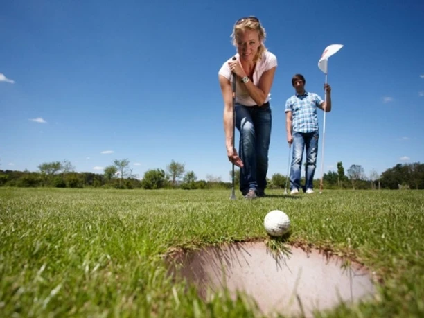 SwinGolf Mehringer Heide Frau beim SwinGolf-Abschlag auf grüner Wiese unter blauem Himmel, Mann mit Flagge im Hintergrund.