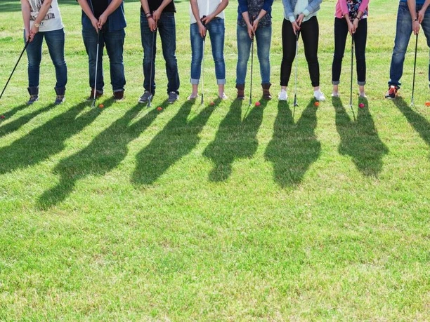 SwinGolf Mehringer Heide Gruppe von Menschen mit Golfschlägern steht auf grüner Wiese, Sonne wirft lange Schatten.