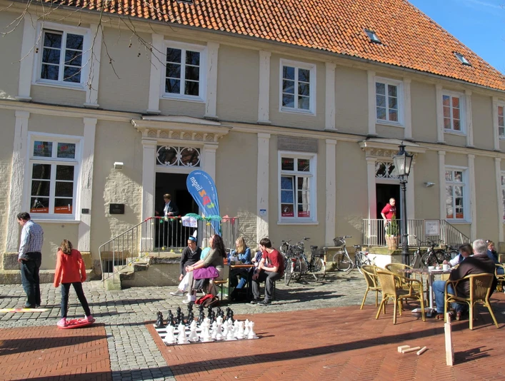 Menschen spielen und sitzen vor einem historischen Gebäude mit rotem Ziegeldach bei sonnigem Wetter.