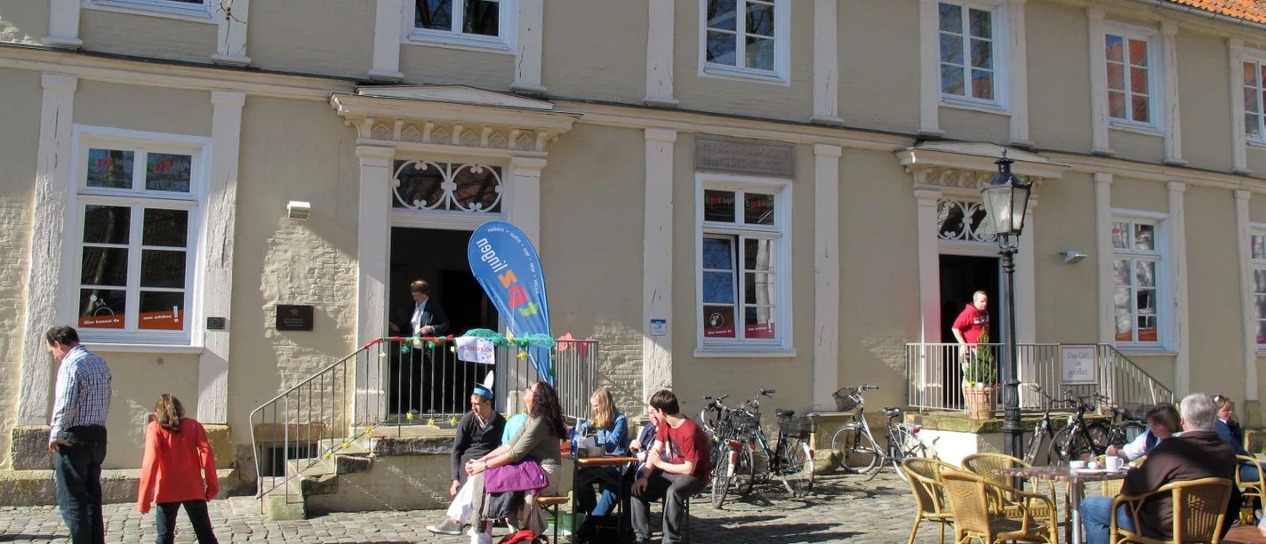 Theaterpädagogisches Zentrum der Emsländischen Landschaft e. V. Menschen spielen und sitzen vor einem historischen Gebäude mit rotem Ziegeldach bei sonnigem Wetter.