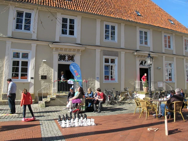 Theaterpädagogisches Zentrum der Emsländischen Landschaft e. V. Menschen spielen und sitzen vor einem historischen Gebäude mit rotem Ziegeldach bei sonnigem Wetter.