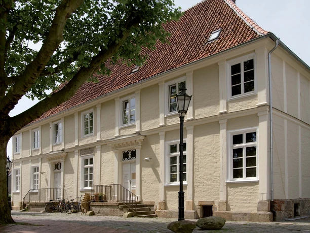 Theaterpädagogisches Zentrum der Emsländischen Landschaft e. V. Zweistöckiges helles Backsteingebäude mit rotem Ziegeldach, Laterne und schattenspendendem Baum davor.