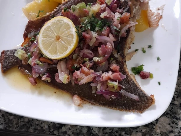 Gebackene Scholle mit Speck, Zwiebeln, Knoblauch, Zitrone und Kartoffeln auf einem weißen Teller.